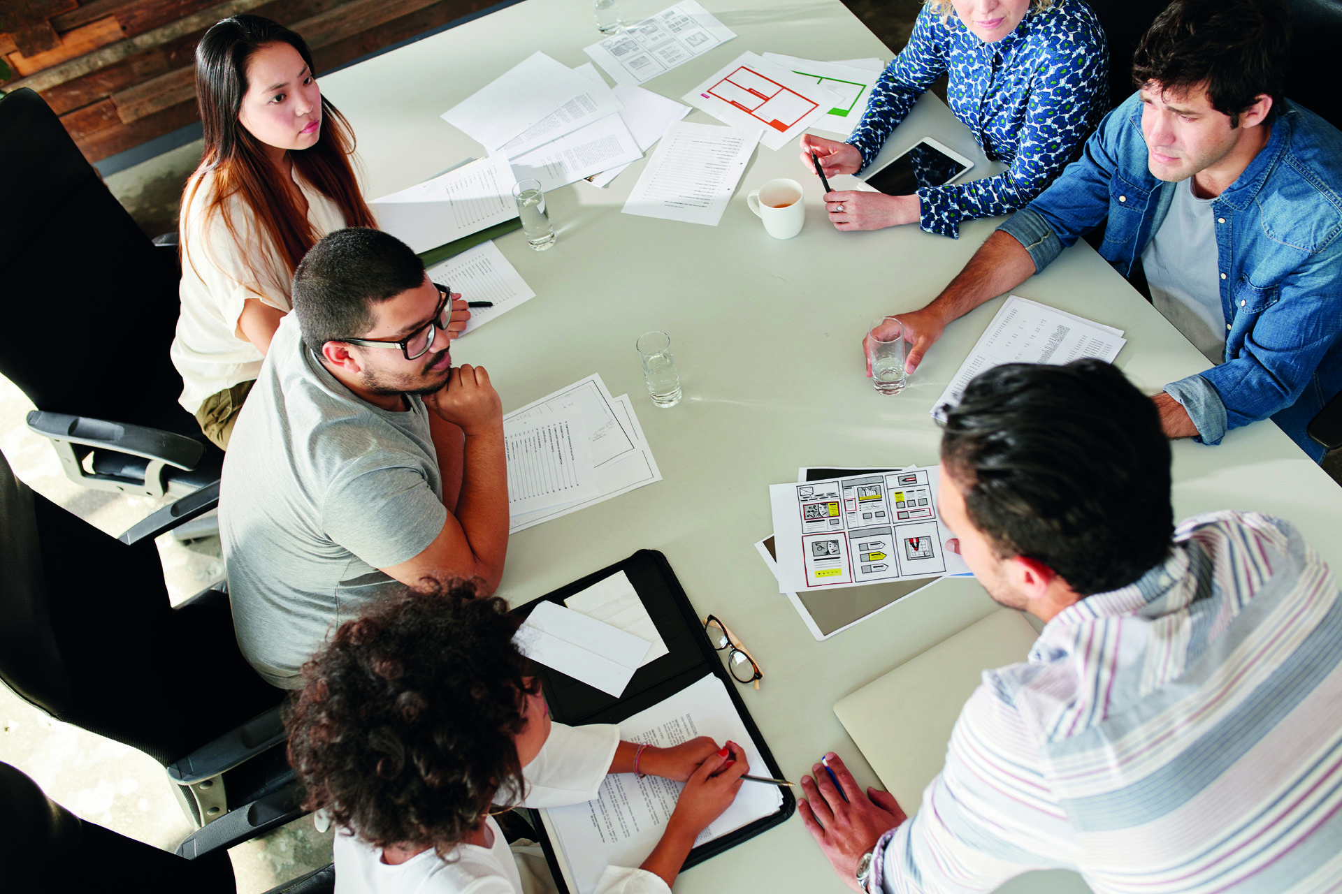 Eine diverse Gruppe junger Leute sitzt gemeinsam am Konferenztisch und bespricht verschiedene Pläne auf Papier.