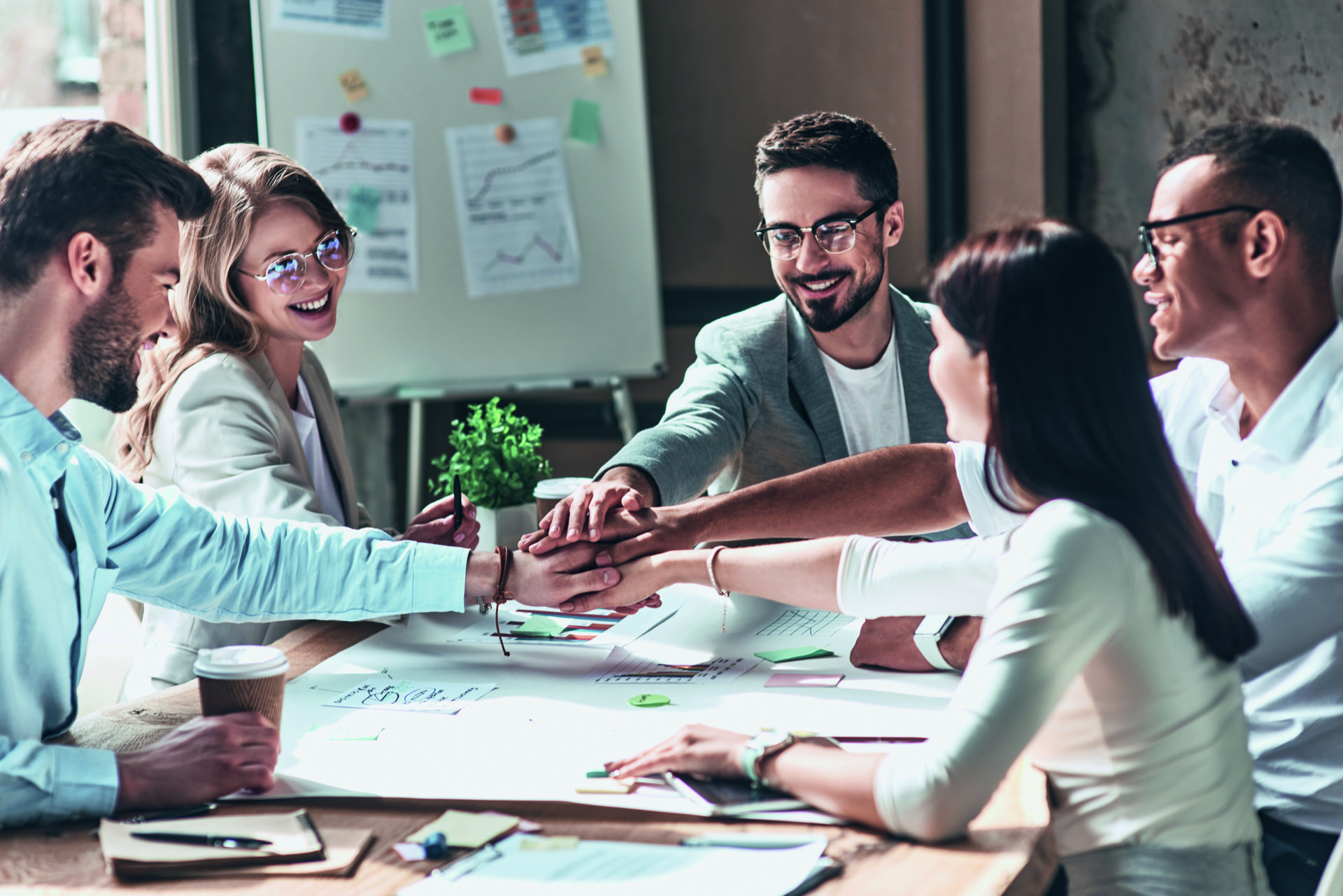 Diverses Team legt die Hand über dem Konferenztisch mit Planungsunterlagen zusammen.
