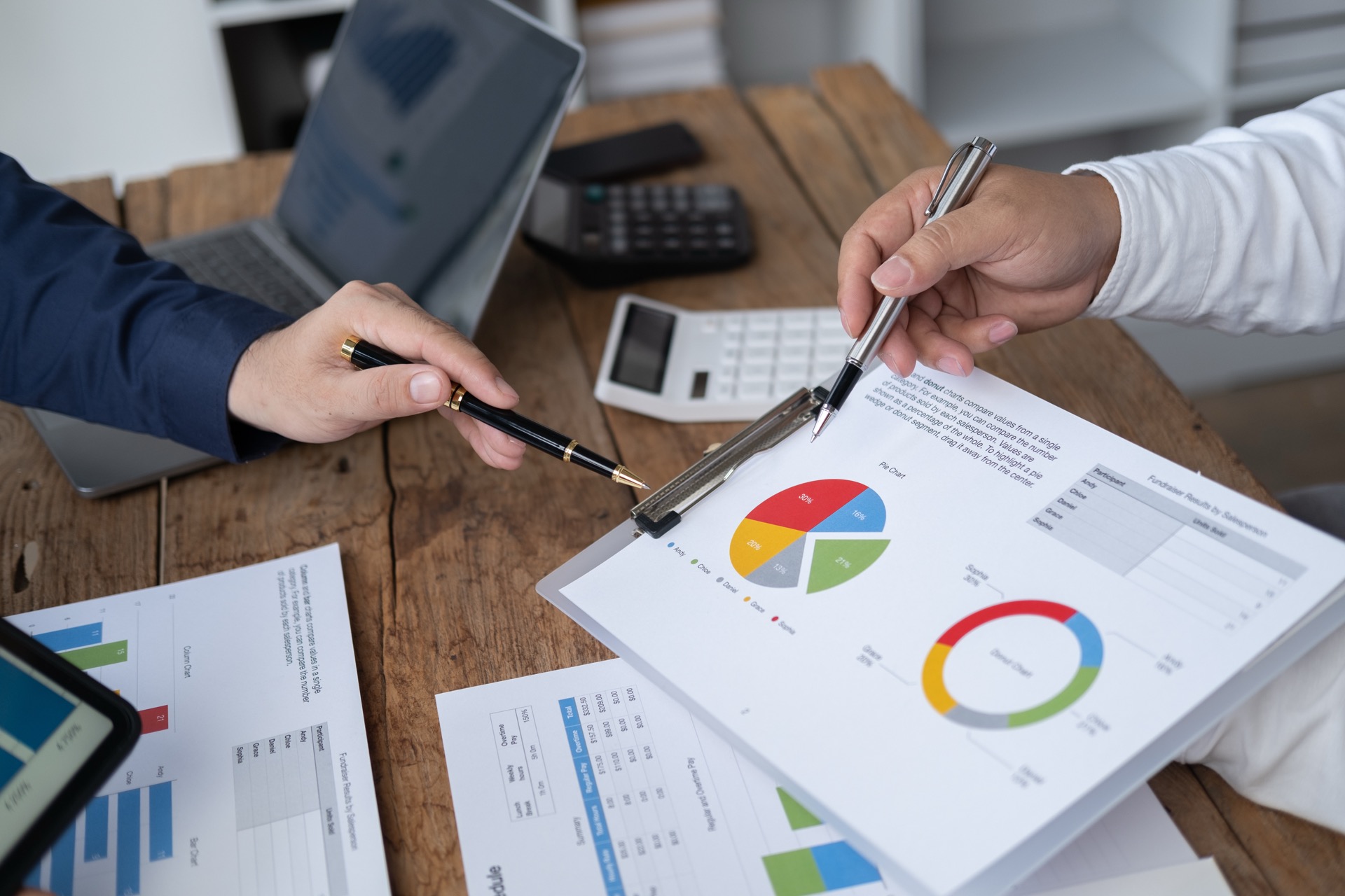 Zwei Hände zeigen auf ein buntes Tortendiagramm. In der Mitte steht ein Schreibtisch mit weiteren Diagrammunterlagen.