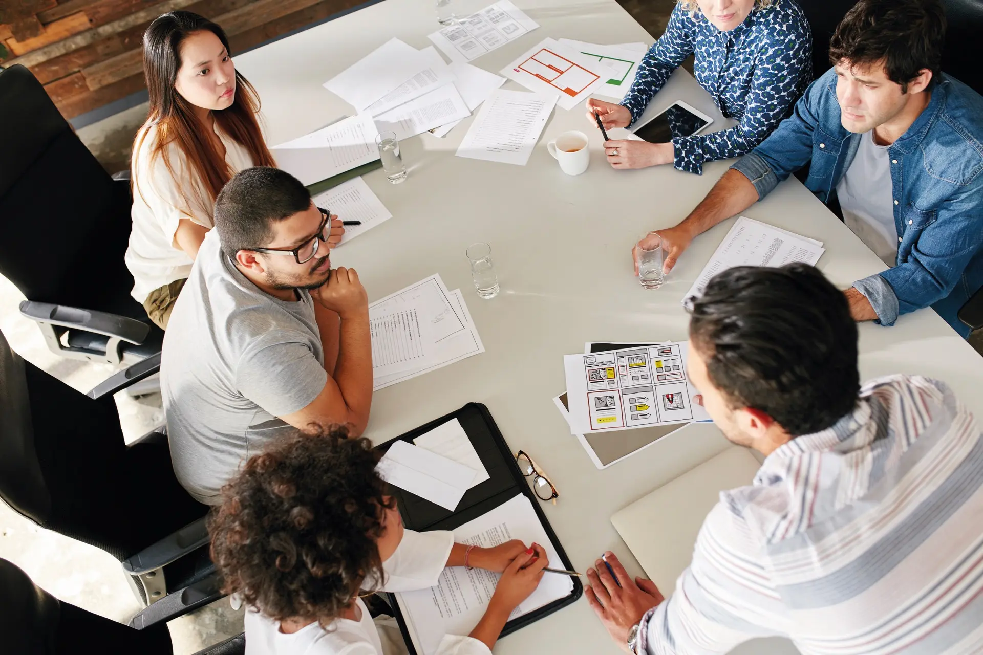 Eine diverse Gruppe junger Leute sitzt gemeinsam am Konferenztisch und bespricht verschiedene Pläne auf Papier.