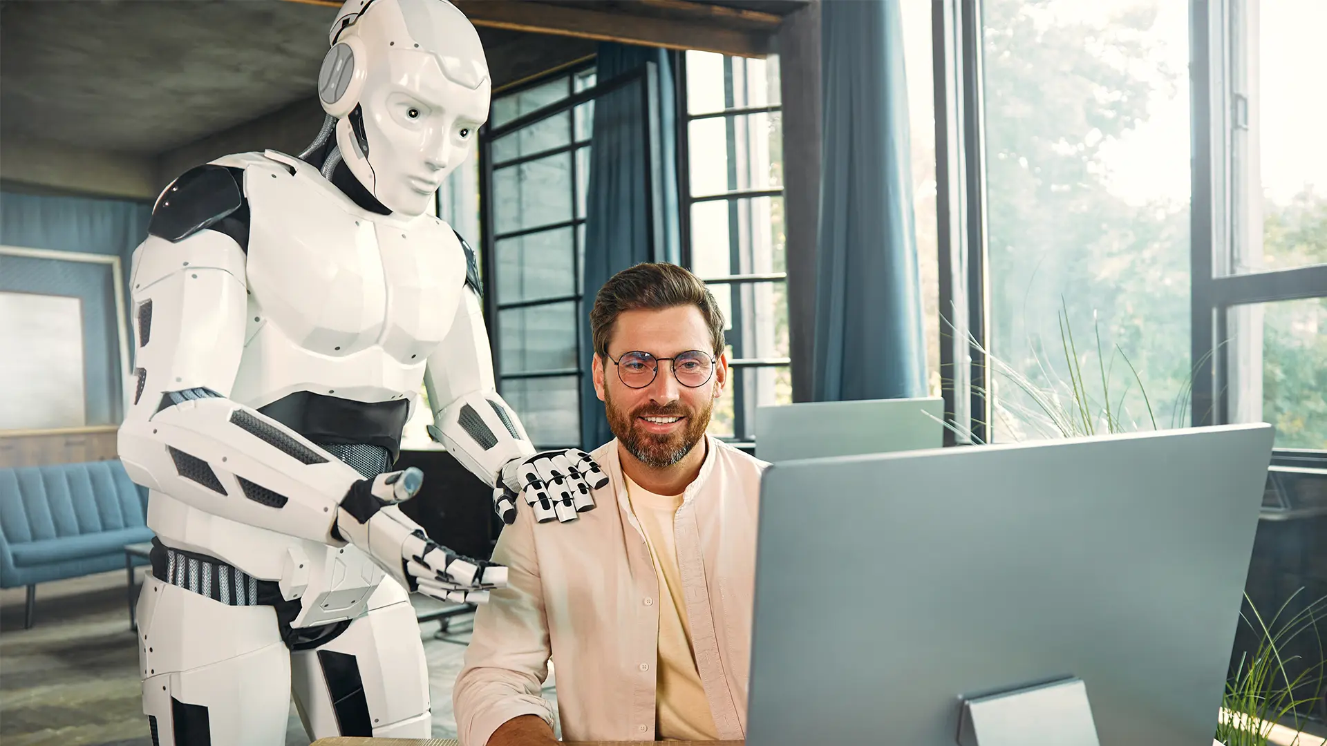 Mann mittleren Alters in beigem T-Shirt und offenem Hemd sitzt am Schreibtisch am Computer und erhält Hilfe von einem neben ihm stehenden humanoiden Roboter, der eine Hand auf der Schulter des Mannes abgelegt hat.