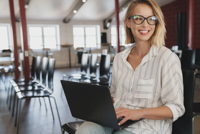 Blonde Frau mit schwarzer, markanter Brille sitzt auf einem Stuhl mit einem Laptop auf dem Schoß in einem großen Raum und schaut rechts schräg aus dem Bild raus.